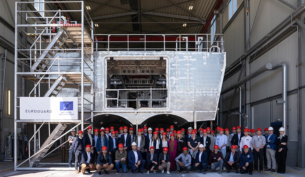 Deelnemers aan de projectbijeenkomst poseren voor het prototype van het schip.